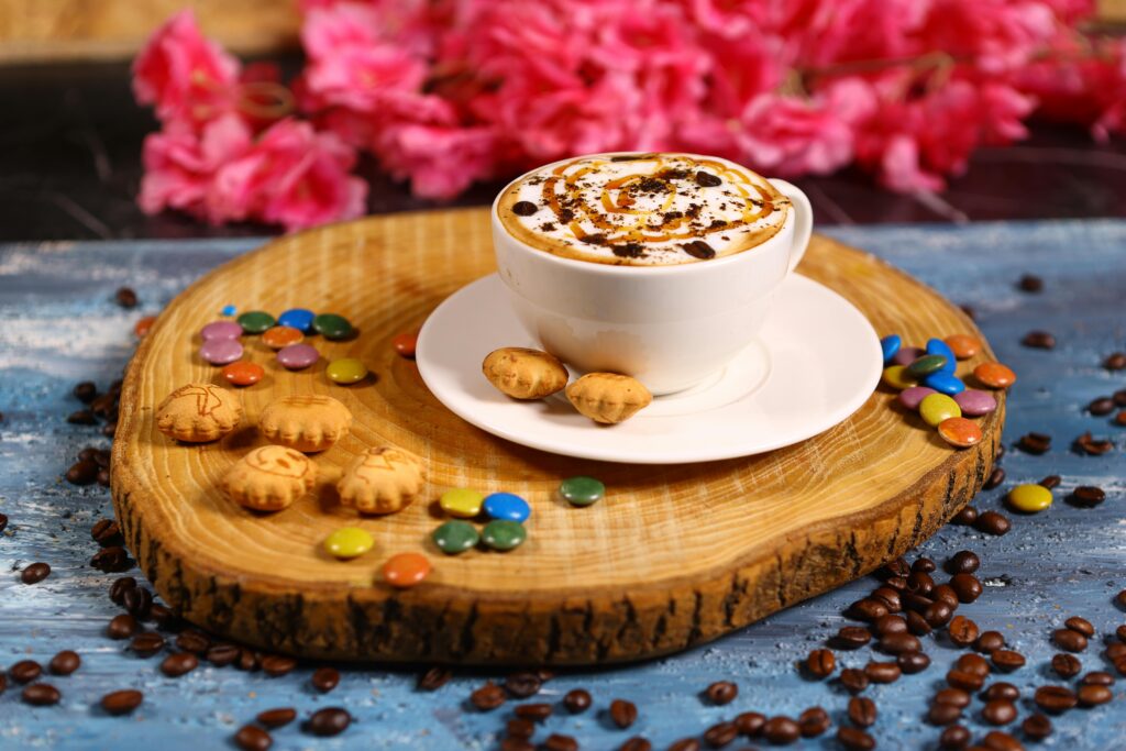 A delicious cappuccino topped with caramel syrup, surrounded by candies and coffee beans, on a wooden slab.