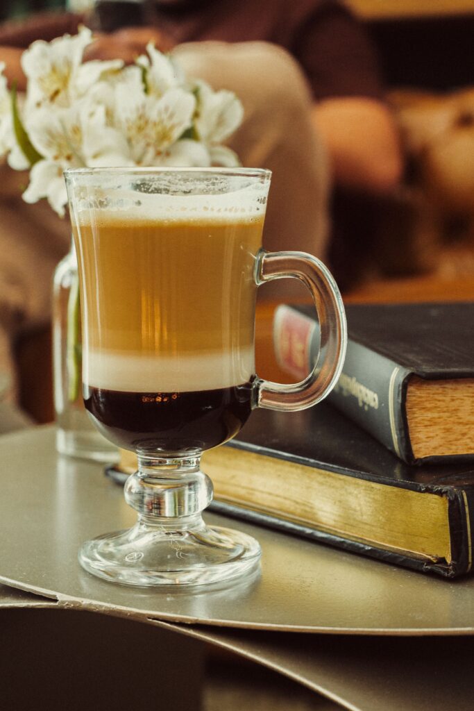 Warm coffee with layered foam in a glass cup beside books and flowers.