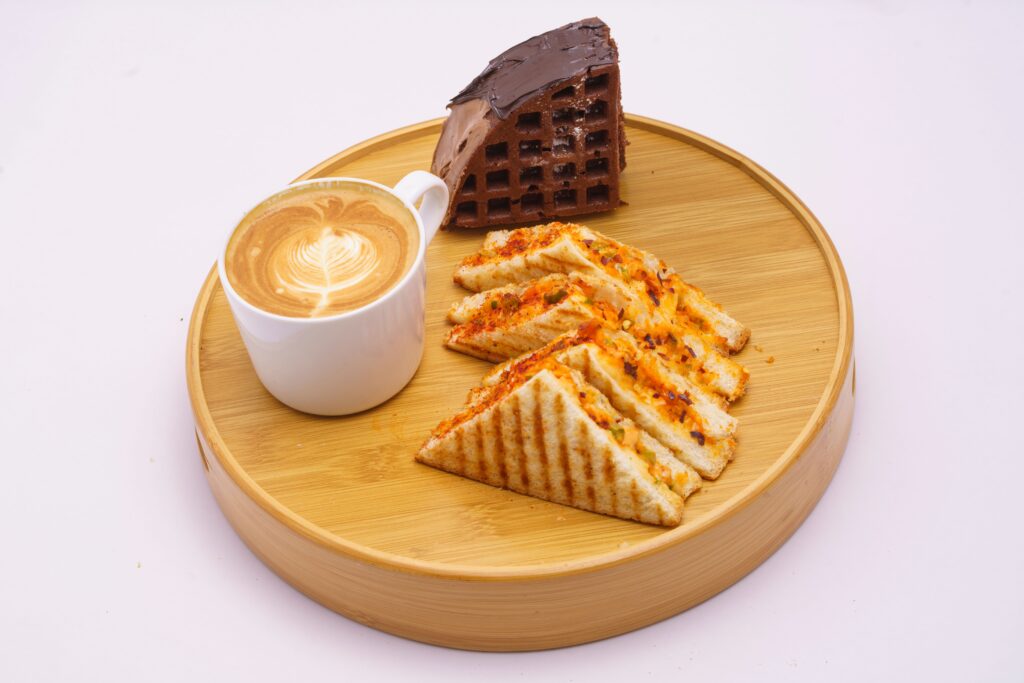 A savory breakfast setup with grilled sandwiches, chocolate cake slice, and latte on a wooden tray.