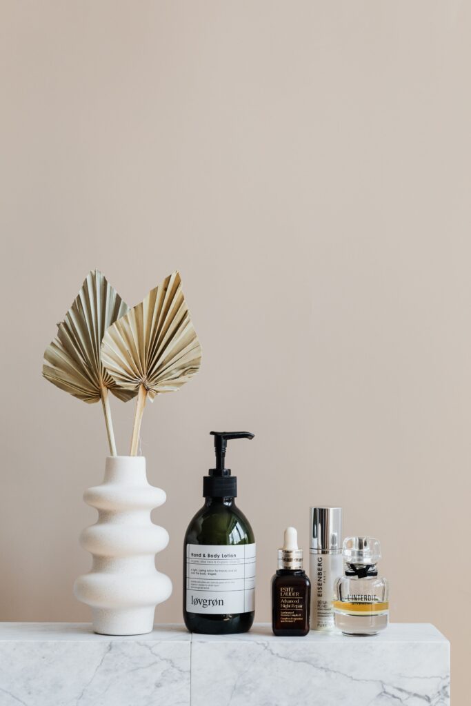 Assorted bottles with cosmetics products placed with creative geometric vase with artificial leaves on white marble shelf against beige wall in modern bathroom interior