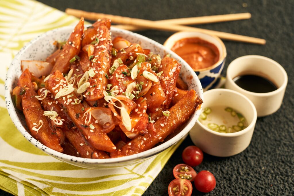 Spicy Korean snack bowl garnished with sesame seeds, accompanied by dipping sauces and fresh cherry tomatoes.