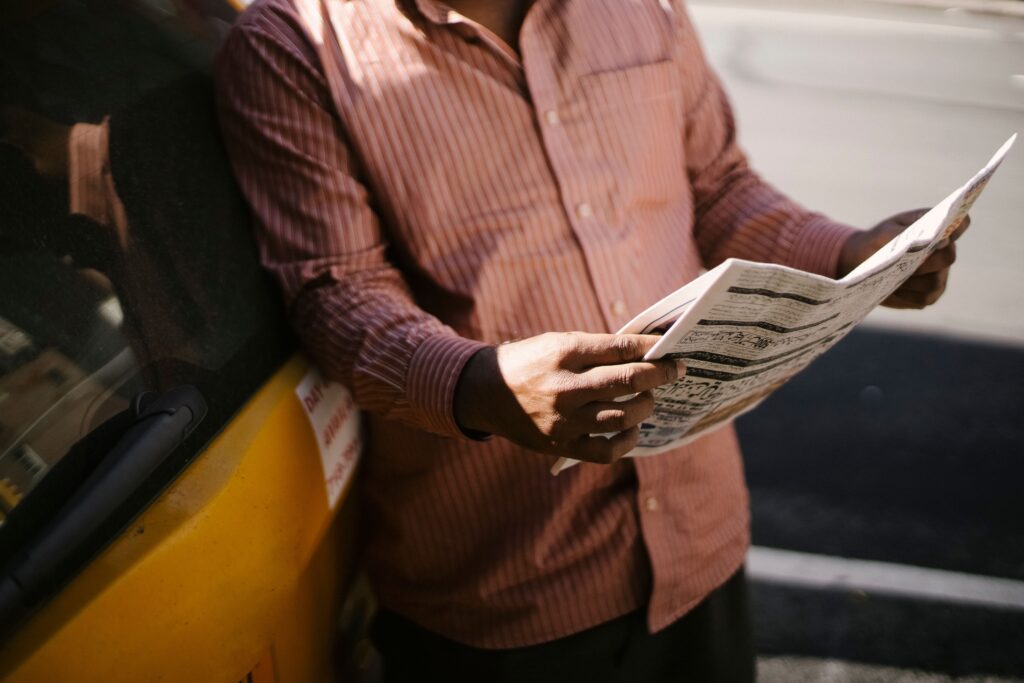 Crop anonymous ethnic cab driver in ornamental shirt standing with newspaper near transport in sunlight