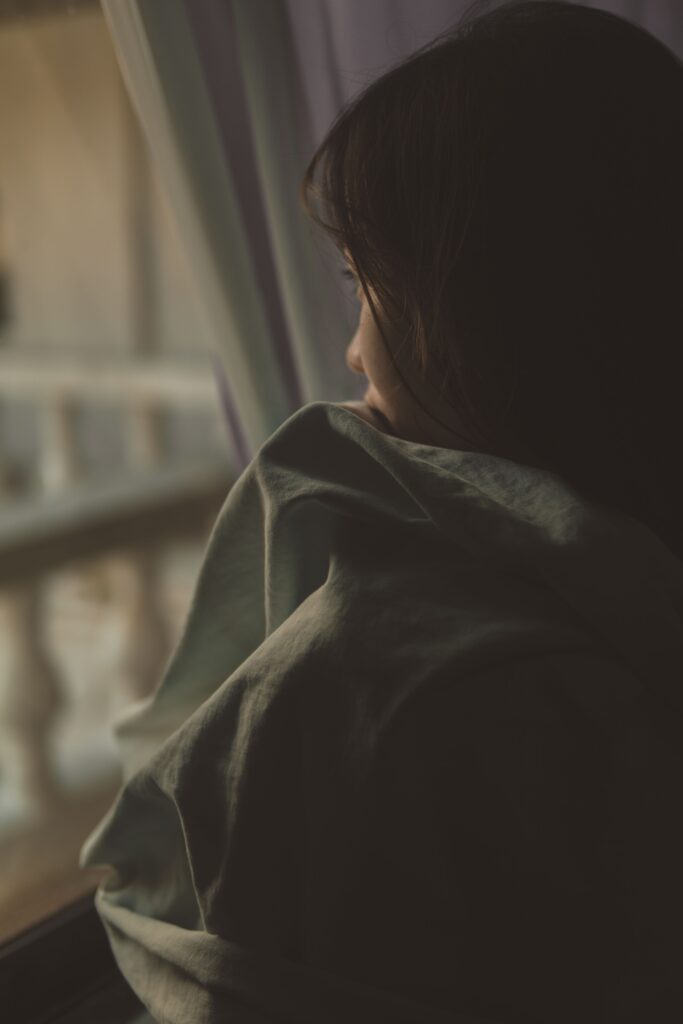 A woman wrapped in a blanket gazing thoughtfully out of a window, captured in a moody ambiance.
