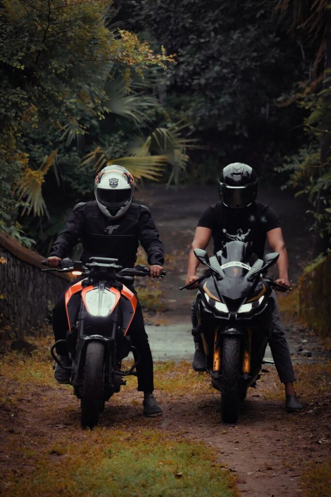 Two motorcyclists in gear on a forest dirt road, surrounded by lush greenery.